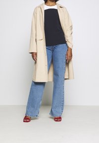 Trench coat beige sopra una maglia blu navy e bianca, abbinato a jeans a zampa d'elefante azzurri e sandali con tacco rossi. Materiale in cotone con cuciture visibili.