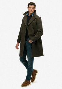 Abrigo de lana verde oscuro con cuello ancho, botones de doble pecho, que se lleva sobre un suéter negro, jeans de mezclilla azules y botas marrones.