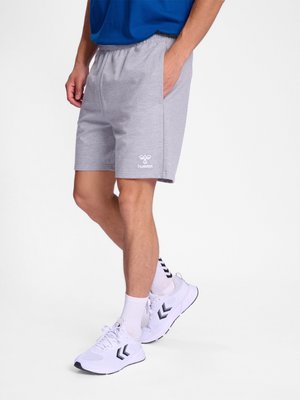 Mann trägt graue hummel-Shorts, weiße Socken mit schwarzen Chevron-Mustern und weiße Sportschuhe mit schwarzen Chevron-Logos, steht mit einer Hand in der Tasche.