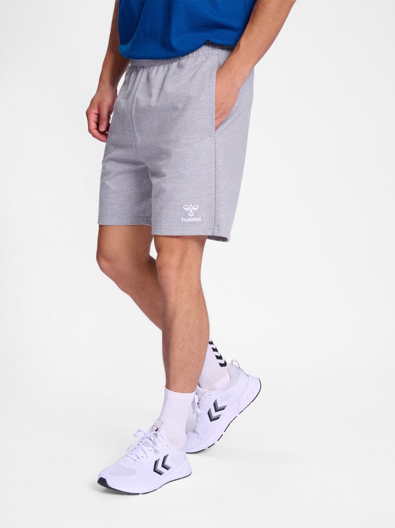 Mann trägt graue hummel-Shorts, weiße Socken mit schwarzen Chevron-Mustern und weiße Sportschuhe mit schwarzen Chevron-Logos, steht mit einer Hand in der Tasche.