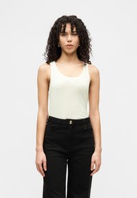 Ανοιχτό πράσινο τοπ με φαρδιές τιράντες και στρογγυλή λαιμόκοψη, σε συνδυασμό με ψηλόμεσο μαύρο παντελόνι με χρυσό κουμπί ως διακοσμητικό στοιχείο.