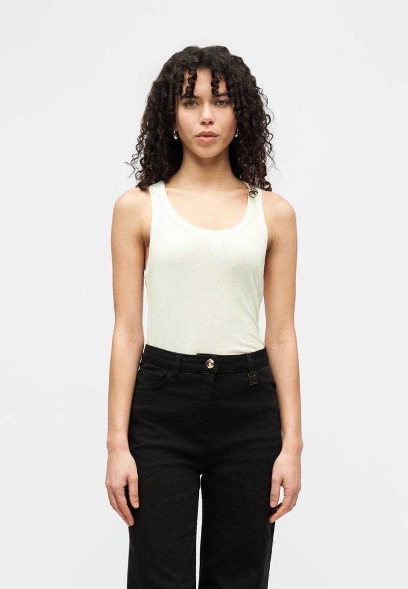 Ανοιχτό πράσινο τοπ με φαρδιές τιράντες και στρογγυλή λαιμόκοψη, σε συνδυασμό με ψηλόμεσο μαύρο παντελόνι με χρυσό κουμπί ως διακοσμητικό στοιχείο.