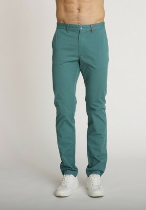 Homme portant un pantalon slim bleu sarcelle et des baskets blanches, debout devant un fond clair uni, les mains dans les poches.