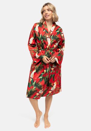 Vrouw staat blootsvoets, draagt een rode knielange jurk met groene bladeren en witte vogelpatronen, handen gevouwen ter hoogte van de taille.