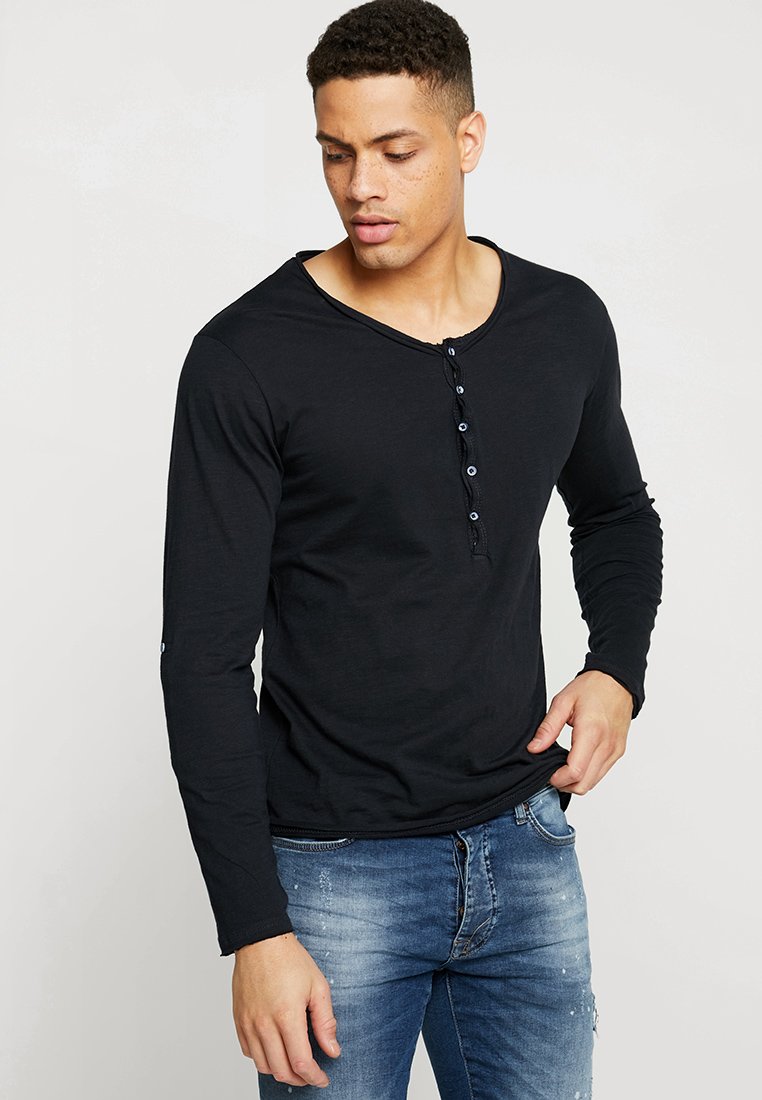 Camiseta negra de manga larga con escote redondeado y tapeta de siete botones. La tela parece suave; el modelo la lleva con unos vaqueros azules.