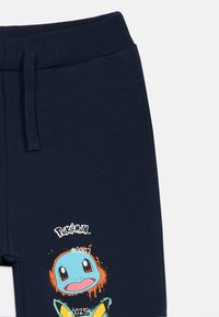 Calças de jogging azul-marinho em mistura de algodão, apresentando uma ilustração de um personagem Pokémon em cores vibrantes e um logotipo acima.