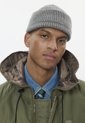 Chaqueta verde con cremallera y un interior brillante, superpuesta sobre una camisa azul clara y una corbata a rayas verdes. Un gorro de punto gris con canalé completa el look.