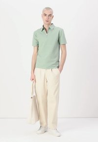 Svetlozelena polo majica s kratkimi rokavi in zadrgo na ovratniku, smetana široke hlače in bele superge, drži bež vrečko.