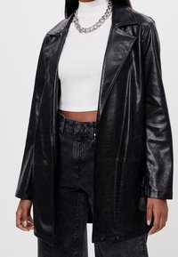Czarna kurtka z sztucznej skóry z klapowym kołnierzem i prostym krojem, z teksturowaną powierzchnią i dwoma przednimi kieszeniami. W zestawie z białą bluzką.