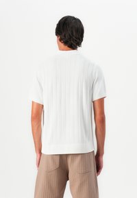 Man with dark hair wearing a white short-sleeve ribbed knit shirt and light brown pants, standing facing away against a white background.