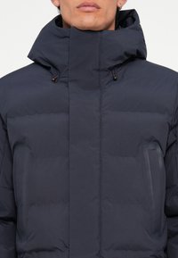 Navy puffer jacket with a high collar, featuring two side pockets and a quilted texture. Closure with drawstring adjustments at the neck.