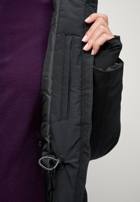 Zwarte jas interieur met een zachte paarse fleece voering, voorzien van een zak, rits en trekkoorddetail. Getextureerde mouwen en schone naden.