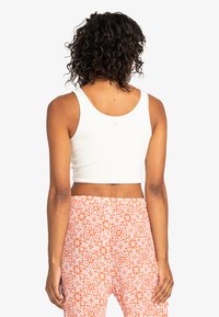 Biely rebrovaný crop top s okrúhlym výstrihom, kombinovaný s oranžovými vzorovanými nohavicami s kvetinovými a geometrickými motívmi.