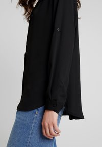 Black long-sleeve blouse with a smooth texture, featuring a buttoned cuff and a slight high-low hem, paired with light blue jeans.