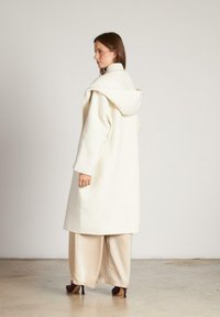 Lungo cappotto bianco con cappuccio, realizzato in tessuto morbido. Presenta spalle scese e una vestibilità rilassata, abbinato a pantaloni larghi color crema e tacchi alti.