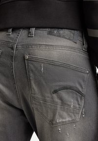 Primer plano de una persona que lleva puestos unos vaqueros de mezclilla gris desgastados, con un bolsillo trasero, detalles de costura y una etiqueta de marca de cuero negro en la cinturilla.