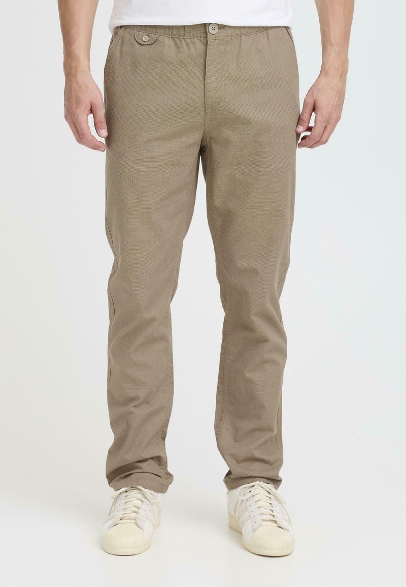Lichtbruine, gestructureerde broek met een relaxte pasvorm, twee voorzakken en knoopsluitingen; gecombineerd met witte sportschoenen.