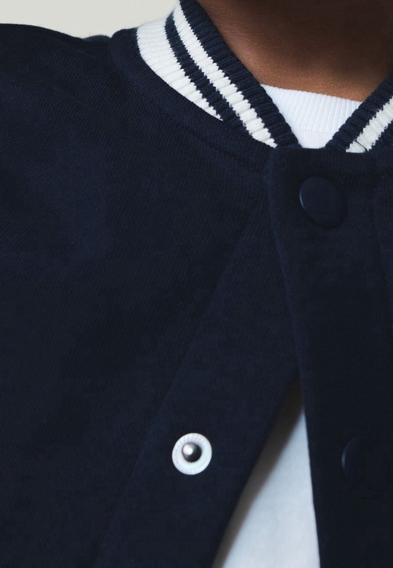 Gros plan d'une personne portant une veste varsity bleu marine avec un col à rayures blanches et des boutons-pression sur une chemise blanche.
