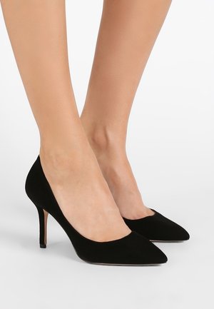 Černé semišové vysoké podpatky se špičatou špičkou, nošené na nohou s tenkými kotníky, zobrazené na jednobílém pozadí.