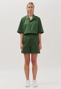 Camicetta verde in cotone a maniche corte e pantaloni coordinati con cintura elastica. Presenta una tasca sul petto e una vestibilità comoda. Sneakers bianche.