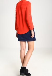 Röd långärmad blus med rundad fåll, tillsammans med en mörk denimkjol. Svarta ankelstövlar fulländar outfiten. Slät tygkänsla.