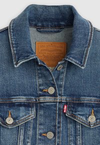 Τζιν μπουφάν με κλασική μπλε απόχρωση, που διαθέτει μυτερό γιακά, μεταλλικά κουμπιά και ετικέτα δερμάτινης μάρκας στο εσωτερικό τη ς περιοχής του λαιμού.