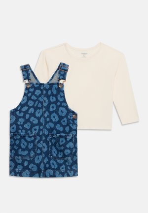 Chemise crème à manches longues associée à une robe salopette en denim bleu avec imprimé léopard foncé et poches avant.