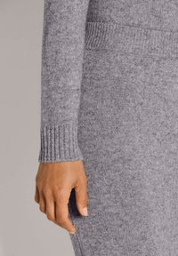 Die Hand entspannt an der Seite, trägt ein hellgraues gestricktes langärmeliges Oberteil und passende strukturierte Hose.
