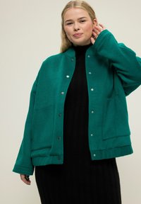 Tyrkysová fleecová bunda se zacvakávacími knoflíky, dvěma předními kapsami a volným střihem. Modelka má pod ní na sobě černé žebrované šaty.