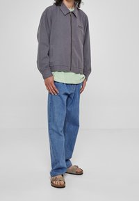 Šedá bunda se zipem vpředu a límcem, vybavená kapsou na hrudi. Kombinovaná se světle zelenou košilí, modrými džínami a béžovými sandály.