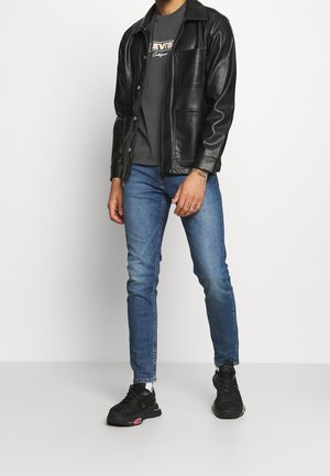 Mand iført sort læderjakke, sort Levi's grafisk t-shirt, blå slim fit jeans, sorte sneakers, stående mod hvid baggrund.