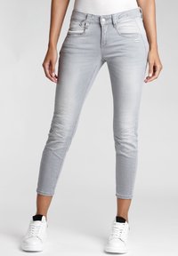 Jeans skinny de mezclilla gris claro con una textura suave, que cuentan con cinco bolsillos, un cierre de botón en la parte delantera y longitud hasta el tobillo. Zapatillas blancas visibles.