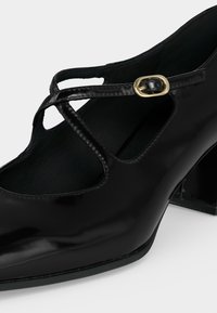 Chaussure Mary Jane en cuir verni noir avec un petit talon carré et une boucle dorée sur des brides croisées.