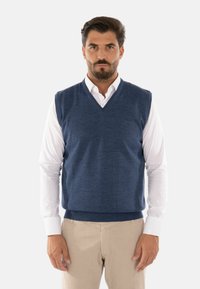 Maglione senza maniche blu navy con scollatura a V, morbido al tatto e con orlo a coste, indossato sopra una camicia bianca con colletto, abbinato a pantaloni chiari.