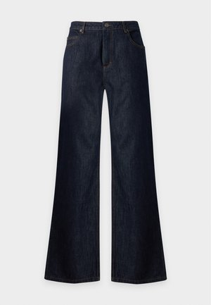 Femme debout portant un pull rose clair avec des poignets côtelés et un pantalon large bleu marine sur un fond blanc uni.