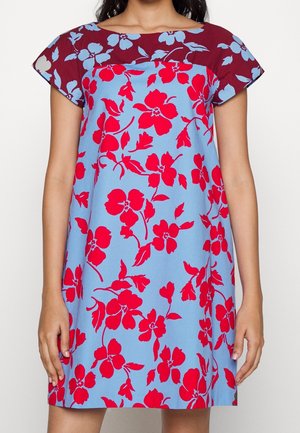 Femme portant une robe à manches courtes avec des motifs floraux rouges sur des sections bleu clair et rouge foncé, debout, bras détendus le long du corps.