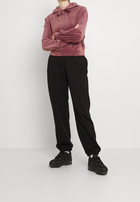 Felpa con cappuccio color rosa antico con cappuccio regolabile, abbinata a pantaloni jogger neri. Le scarpe sono sneakers nere con suole spesse.