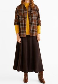 Chemise écossaise marron avec poches avant, pull en tricot jaune et jupe maxi marron. Des bottes à talons marron complètent la tenue.