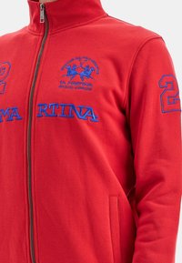 Giacca rossa con zip in tessuto morbido e texturizzato, con logo del marchio ricamato in blu, grafica di un giocatore di polo e dettagli con numero sulla manica.