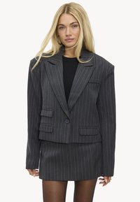 Mujer rubia con blazer de rayas gris oscuro de gran tamaño y falda a juego, con camiseta negra, posando contra un fondo liso.