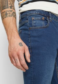 Σκούρο μπλε τζιν παντελόνι με στενή γραμμή, αντίθετη ραφή και κανονικές τσέπες. Χέρι με τατουάζ ορατό στη μία πλευρά.