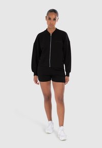 Felpa nera con zip, polsini e orlo a coste, abbinata a pantaloni corti neri. Il modello indossa sneakers bianche. Design semplice e di vestibilità rilassata.