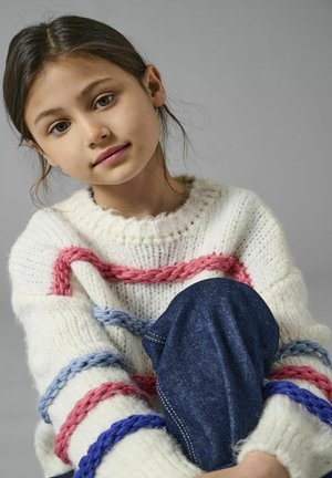 Pull en tricot blanc avec des accents tressés rouge, bleu et rose. Jean en denim bleu. Gros plan d'une jeune fille assise, avec un fond neutre.
