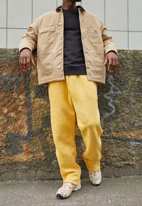 Tan canvasjakke med store lommer, sort sweatshirt under, gule sweatpants og multicolor sneakers. Tekstureret stenbaggrund.