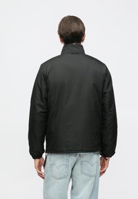 Casaco acolchoado preto com gola alta, punhos elásticos e textura suave, visto por trás. Usado com jeans de ganga claro.