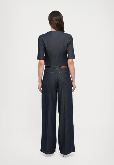 Giubbotto in denim scuro con top corto e pantaloni a gamba larga. Il tessuto ha una consistenza liscia, accentuata da cuciture a contrasto e tasche.
