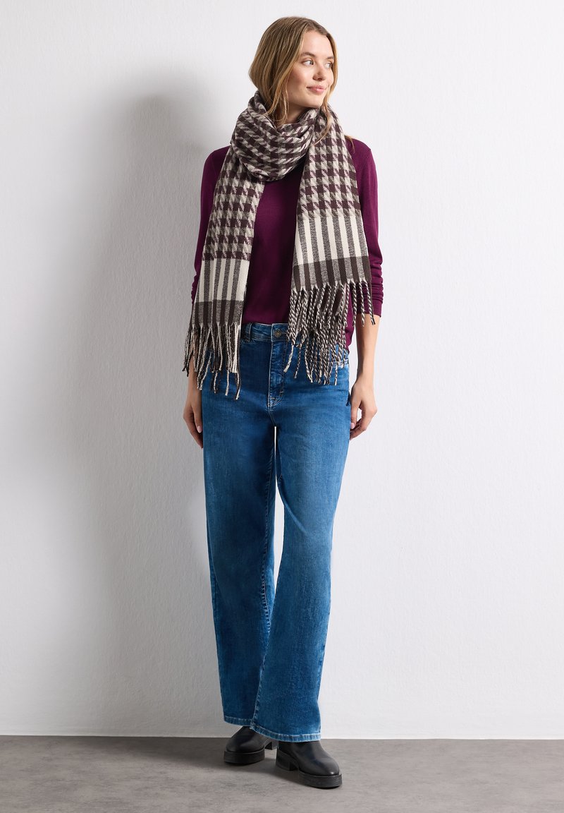 Femme portant un jean bleu, des bottes noires, un haut bordeaux et une grande écharpe à carreaux, debout contre un mur blanc, regardant vers la droite.