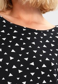 Haut noir orné d'un motif de triangles blancs dans diverses orientations. Le tissu semble lisse avec un décolleté décontracté.