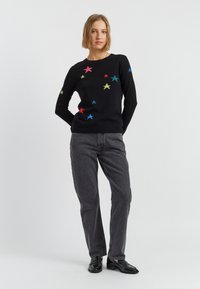 Zwarte gebreide trui met kleurrijke ster-applicaties in rood, blauw, geel en groen. Gecombineerd met grijze jeans en zwarte schoenen.