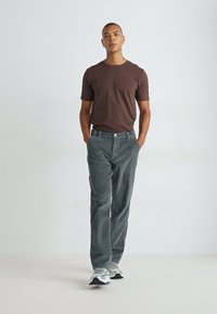 Brown short-sleeve shirt, gray corduroy pants, and athletic shoes. Model stands with hands in pockets against a light background.